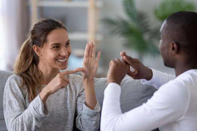 Smiling mixed ethnicity couple or interracial friends talking with sign finger hand language, happy two deaf and mute hearing impaired people communicating at home sit on sofa showing hand gestures