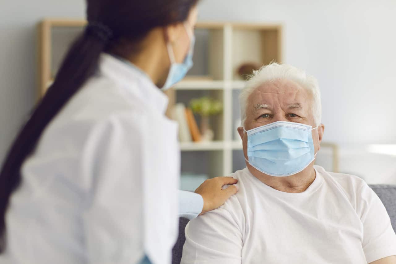 People in protective masks. Woman doctor visits the elderly man and provides him with support and talks to him during the quarantine. Nurse visits a man who is at risk.